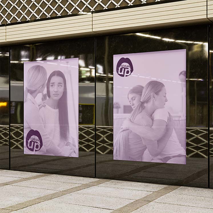 City environement where there is two large purple posters with a logo at the bottom featuring an open mouth saying speak up. They both have a photo of two women in an emotional conversation.